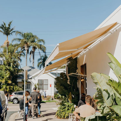 Customers relax outside a white building under a beige awning, sunny day.